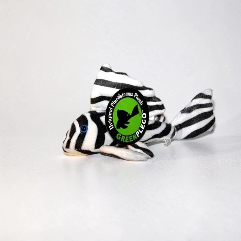 Ein Stofftier in Form eines gestreiften Saugwels (Pleco) auf weißem Grund. Es hat schwarze und weiße Querstreifen, ein kleines blaues Auge und eine grüne runde Plakette mit "Original Plecostomus Plush GREENPLECO".