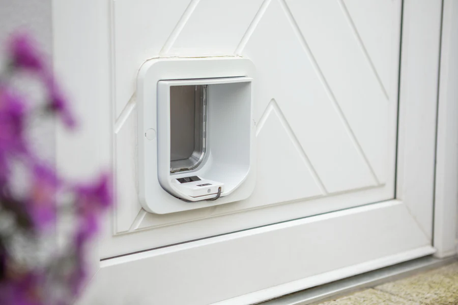 Weiße Katzenklappe in weißer Tür mit diagonalen Zierleisten. Die Klappe ist offen und zeigt den Tunnel dahinter. Unterhalb der Öffnung ein kleines weißes Gerät mit Kette, vermutlich ein Chip-Leser. Links vorn unscharf violette Blüten.