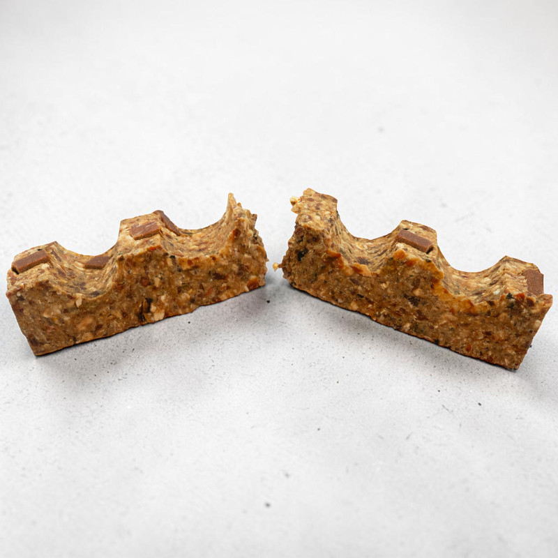 Una barretta naturale, spezzata in due metà, giace su uno sfondo grigio chiaro. La superficie presenta numerose noci, semi e pezzetti di cioccolato fondente. Il bordo superiore di entrambe le metà è ondulato, come piccole collinette.