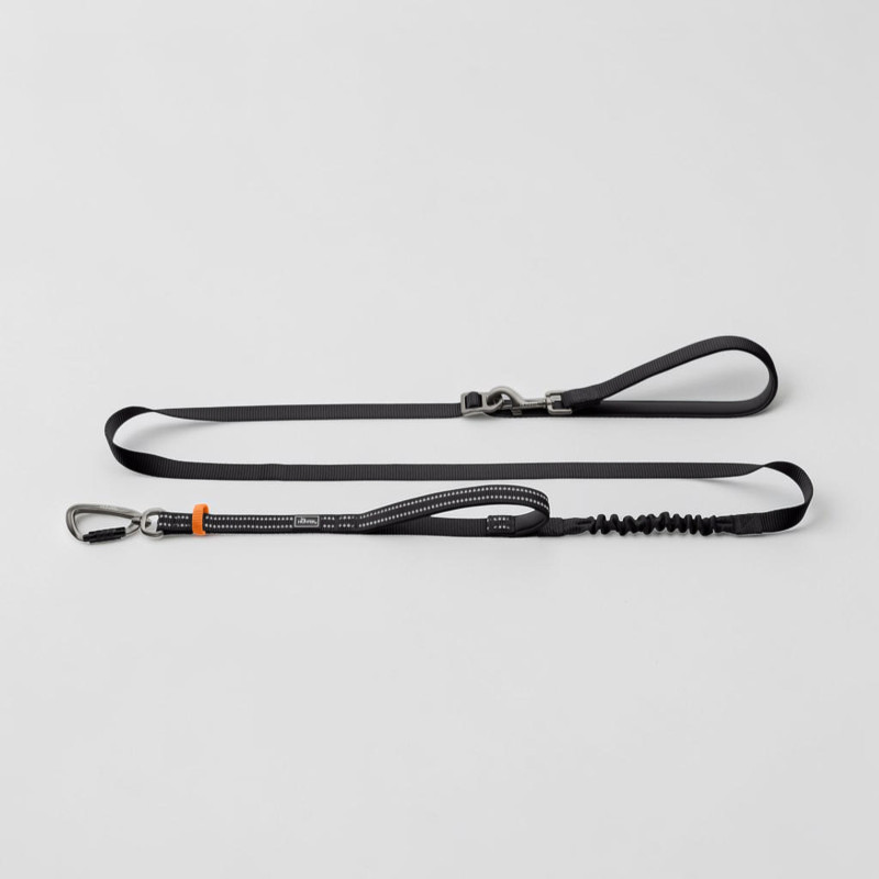 Laisse pour chien noire avec éléments réfléchissants, déployée. Équipée d'un mousqueton en aluminium rehaussé d'une touche orange et d'un amortisseur de chocs intégré. Deux poignées rembourrées, dont une servant de poignée de retenue. Réglable.