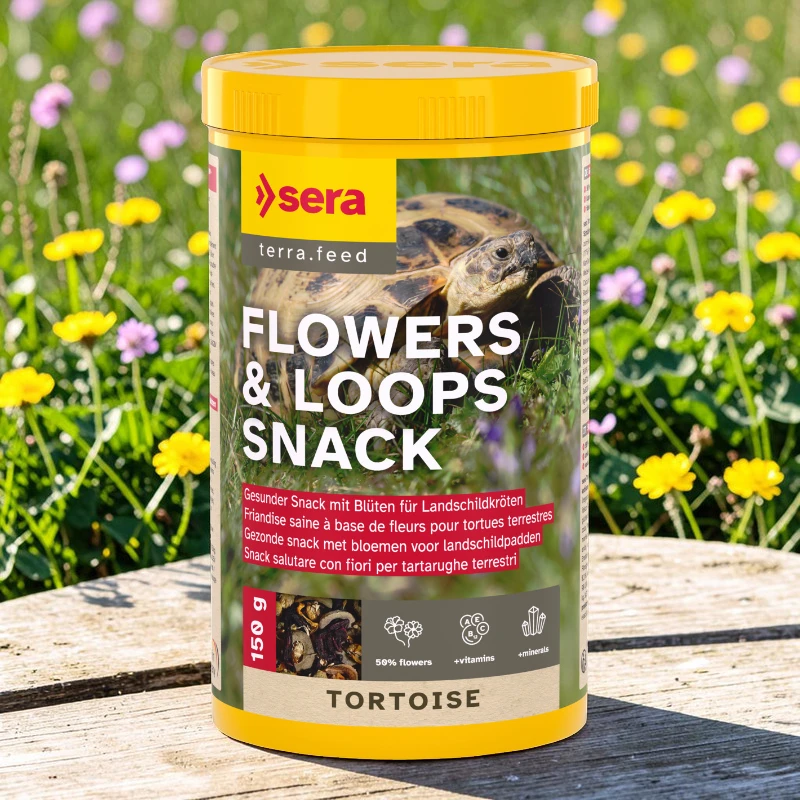 Pot jaune de snack pour tortues Sera « Flowers & Loops Nature » sur support en bois. Étiquette: tortue, fleurs. Texte: « FLOWERS & LOOPS SNACK », « 50 % fleurs », « + vit. », « + min. », 150 g.