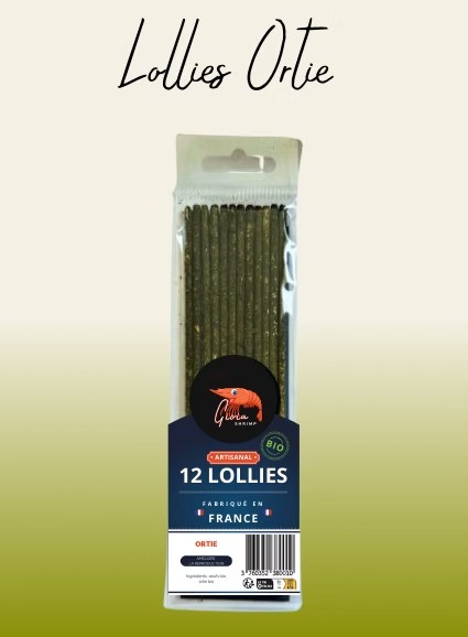 Transparente Packung mit 12 dunkelgrünen, länglichen Garnelen-Lollies. Etikett: Gloria Shrimp Logo, „12 Lollies ARTISANAL BIO FABRIQUÉ EN FRANCE“, „ORTIE“ (Brennnessel) zur Reproduktionsförderung. Hintergrund: Verlauf hellbeige–olivgrün.