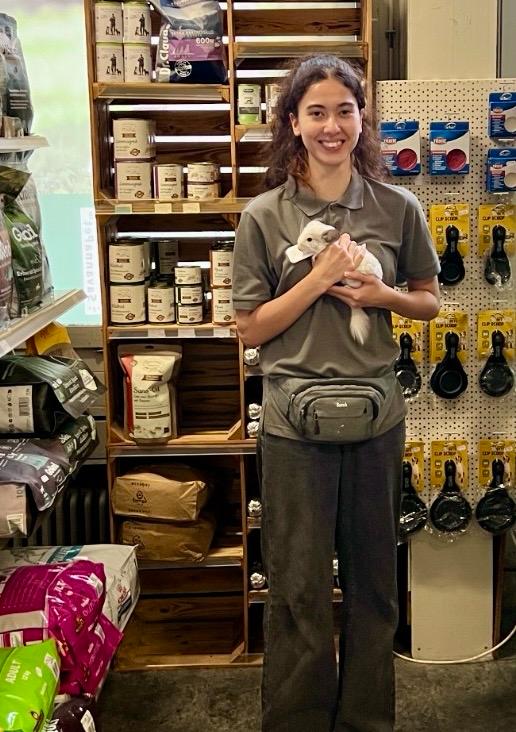 Una giovane donna sorridente dai capelli scuri e ricci tiene delicatamente tra le braccia un piccolo cincillà bianco e soffice. Indossa una polo grigia e un marsupio. Dietro di lei, scaffali con mangimi e accessori per animali nel negozio di animali.