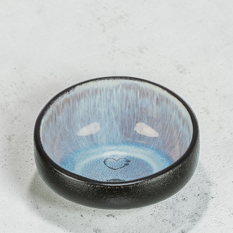 Kleine runde Keramikschale mit rauer, dunkelgrauer Außenseite und glänzender, irisierender Innen-Glasur von Weiß bis Hellblau. Am Boden eine zarte Herz-Silhouette mit eingraviertem Pfotenabdruck. Auf hellgrauem, strukturiertem Untergrund.