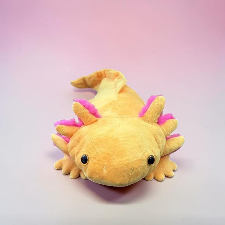 Ein goldgelbes Plüsch-Axolotl mit leuchtend pinken Kiemen liegt mittig auf einer sanft rosafarbenen Oberfläche. Große, runde schwarze Knopfaugen blicken nach vorne. Weicher Stoffkörper mit kleinen Gliedmaßen und gewelltem Schwanz.