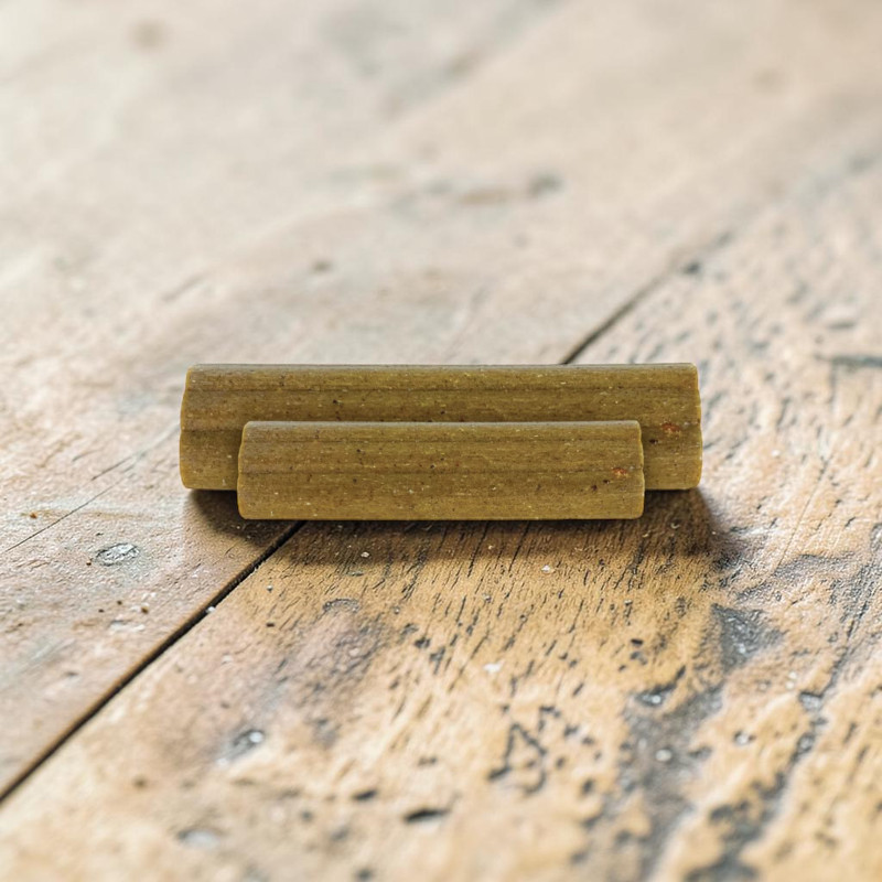 Zwei parallel liegende, zylindrische Kausnacks in bräunlich-grün liegen auf hellbrauner Holzoberfläche. Der obere Riegel ist etwas länger. Beide haben eine leicht unregelmäßige Oberfläche; das Holz zeigt Maserung und Abnutzung.
