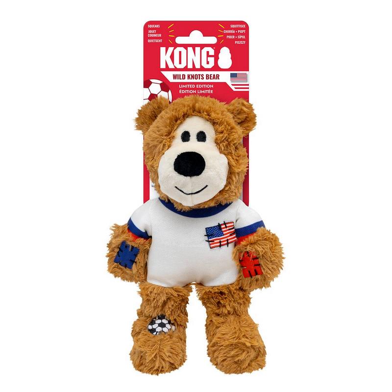 Giocattolo per cani a forma di orso marrone e morbido, in piedi, con maglia bianca con polsini blu e rossi. Sul petto è cucita una bandiera USA. Toppe blu/rosse sulle braccia, pallone da calcio sul piede sinistro. Occhi e naso neri su muso bianco.