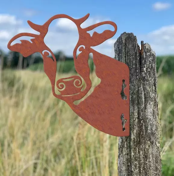 A rusty cow animal silhouette is attached to a wooden post. Grass and a blue sky can be seen in the background.