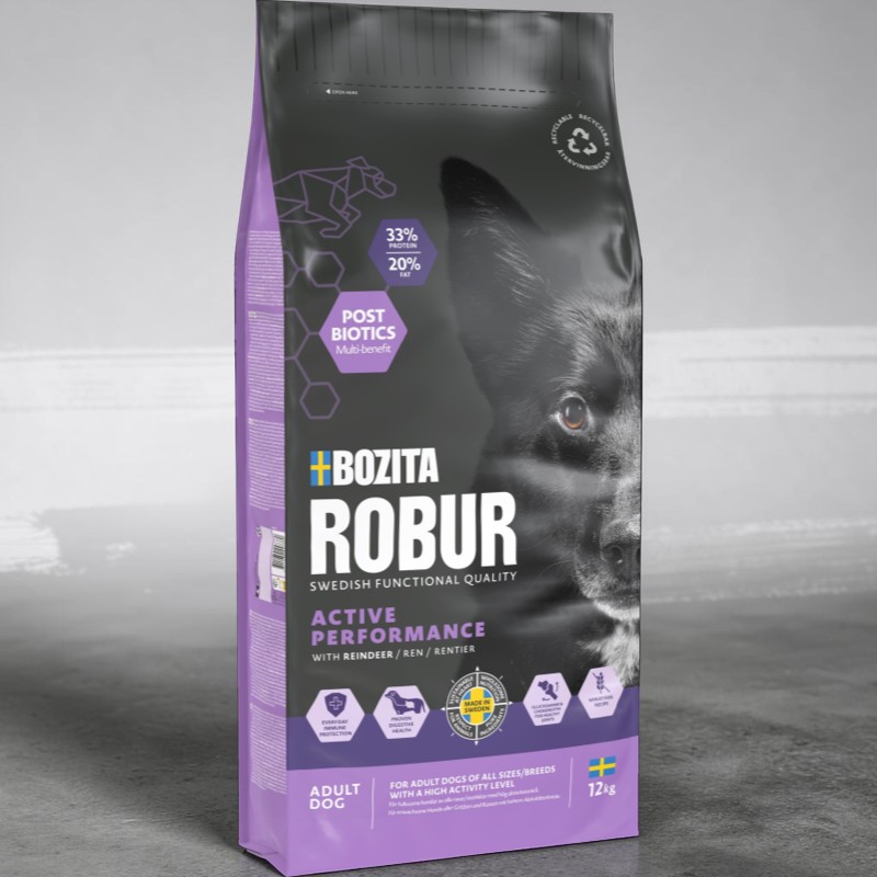 Große dunkelgraue Robur-Hundefutterpackung mit lila Seitenstreifen. Gesicht eines schwarzen Hundes blickt in die Kamera. Bewirbt „Active Performance“ mit Rentierfleisch, 33% Protein, 20% Fett, 12 kg. Post Biotics Multi-benefit.
