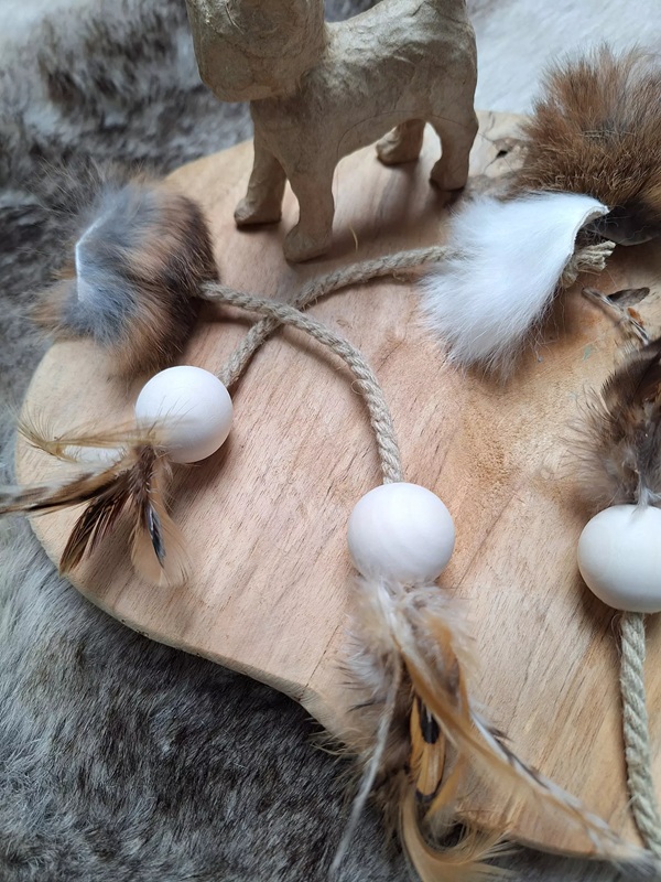 Holzbrett mit Papierhundfigur, daran befestigt Seile mit Holzperlen, Federn und Fellpuscheln. Pelziger Hintergrund.