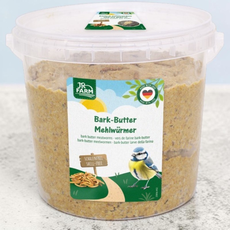 Transparent plastic bucket containing a golden-brown mixture of bark butter and mealworms for birds. Label: a blue tit on a path leading to shell-free mealworms, against a sunny sky. At the top: ‘JR Farm Bark Butter Mealworms’; lid: easy to use.