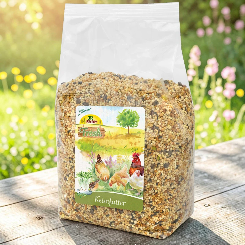 Transparente Plastiktüte mit JR Farm Keimfutter für Hühner, gefüllt mit kleinen Samen. Auf dem Etikett Hennen und Hahn in ländlicher Szene beim Picken an Keimlingen. Tüte auf Holz, unscharfer blühender Garten hinten.