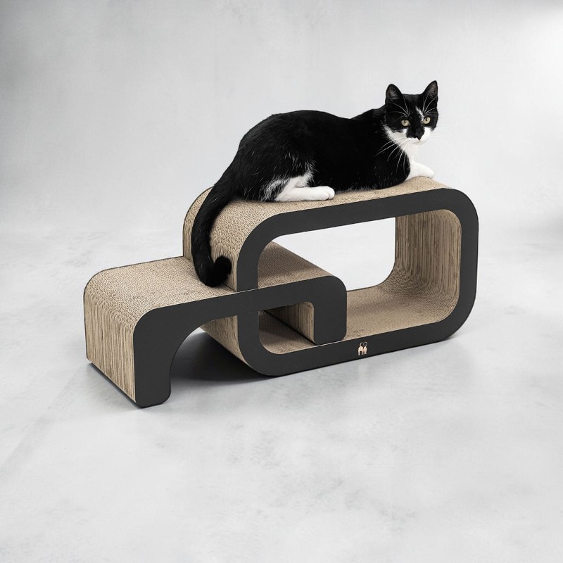 Un gatto bianco e nero è sdraiato su un moderno tiragraffi a due livelli realizzato in cartone ondulato con cornici nere. Guarda attentamente davanti a sé, le zampe bianche e il mento risaltano mentre riposa rilassato in cima.