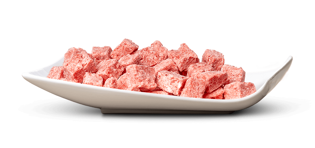 Eine weiße, flache Schale ist gefüllt mit vielen kleinen, unregelmäßigen, rosafarbenen Stücken. Die würfelartigen Teile wirken leicht körnig, wie gefriergetrocknete Früchte oder Süßigkeiten. Hintergrund weiß.