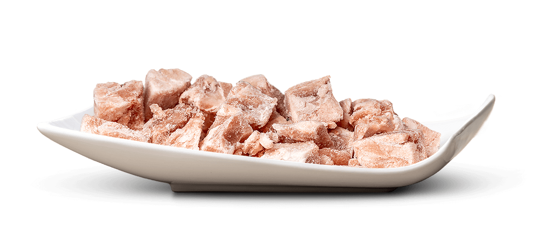 A white, curved ceramic bowl containing numerous irregularly shaped, pale pink to brownish cubes of meat. The frozen pieces are covered in a thin layer of ice and have a matt, frosty appearance. They lie close together.