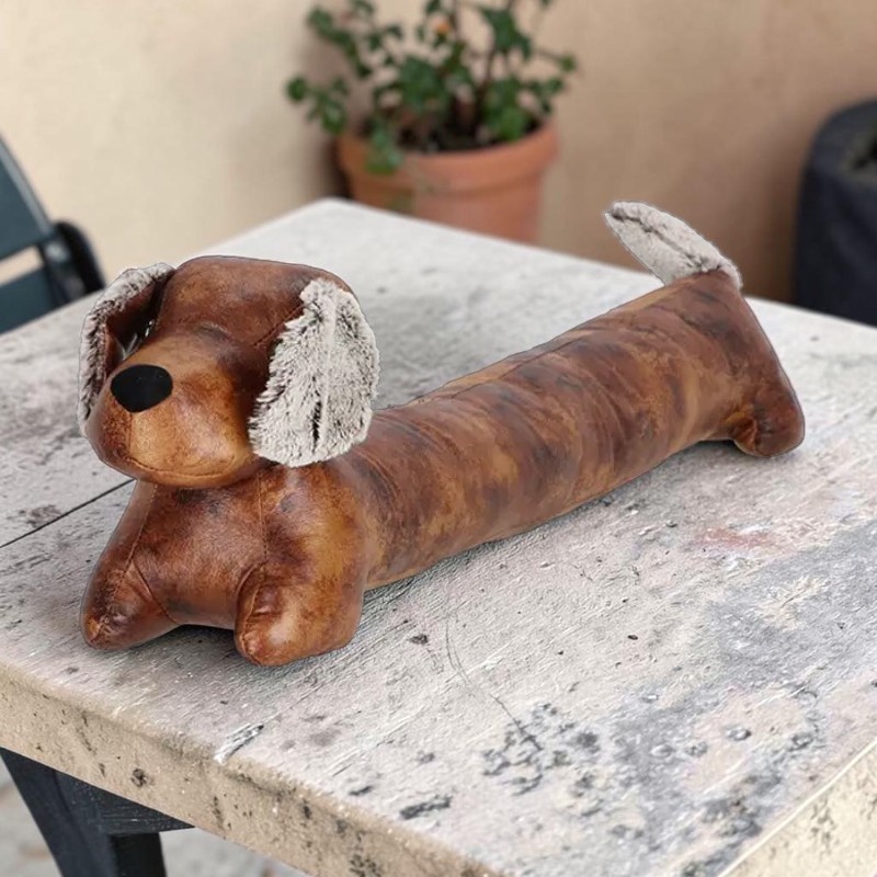 Ein brauner, gepolsterter Dackel-Türstopper mit langem Körper liegt auf einem abgenutzten, hellgrauen Tisch. Er hat flauschige, hellgraue Ohren und eine schwarze Nase. Im unscharfen Hintergrund steht eine grüne Topfpflanze.