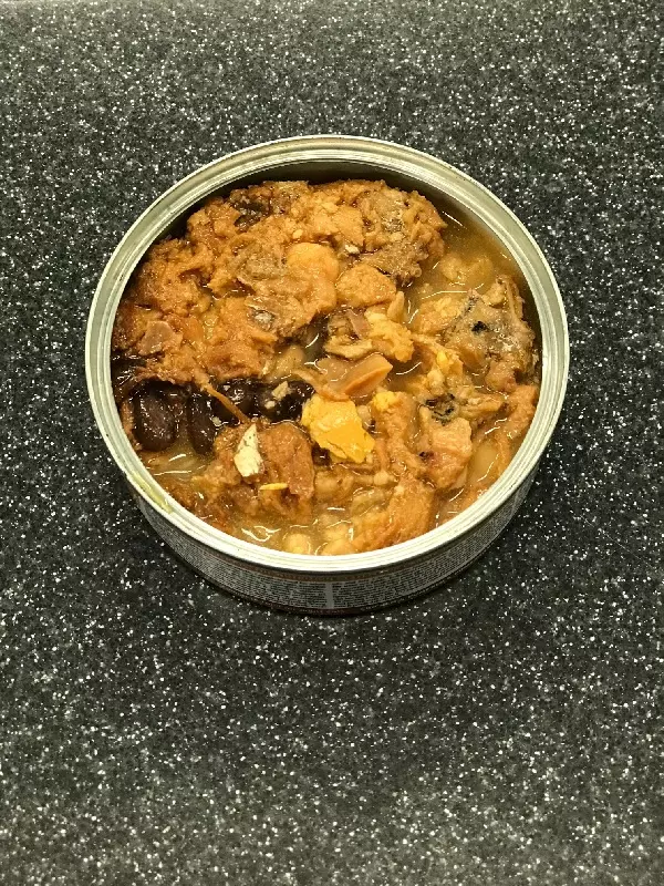 An open tin of animal food, consisting of pieces of meat and gravy, stands on a mottled, dark surface.