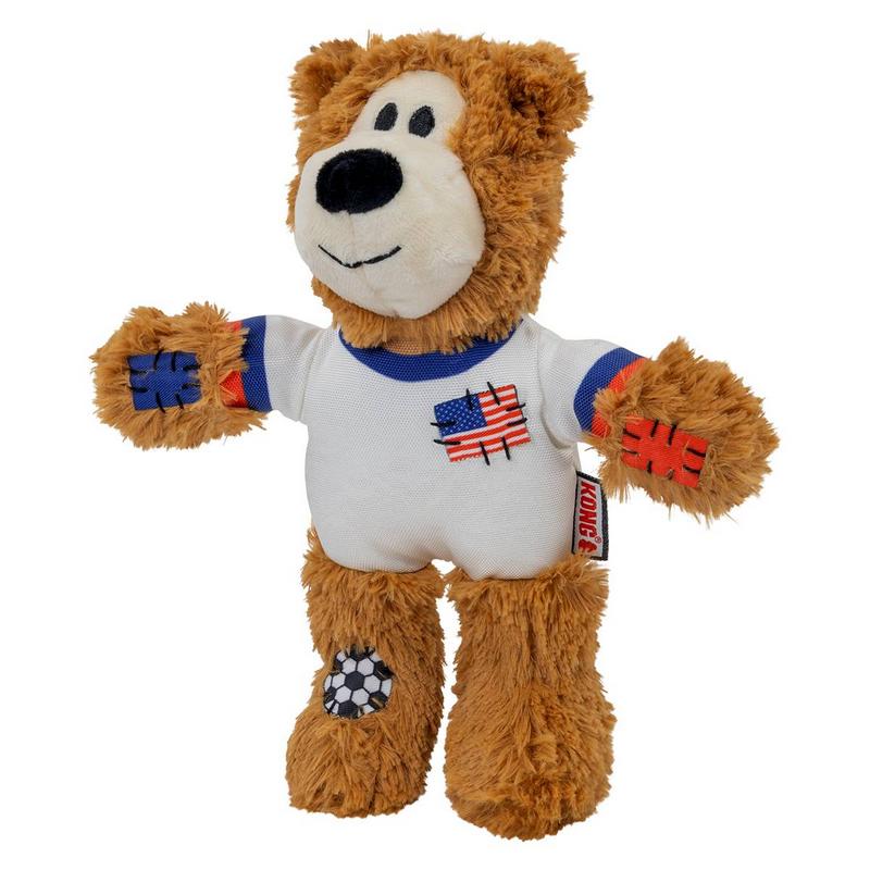 Morbido orsacchiotto di peluche marrone su sfondo bianco. Indossa una maglietta bianca con colletto e polsini blu, patch bandiera USA sul petto. Zampe e un piede con patch blu e arancioni a forma di pallone da calcio. Espressione simpatica.