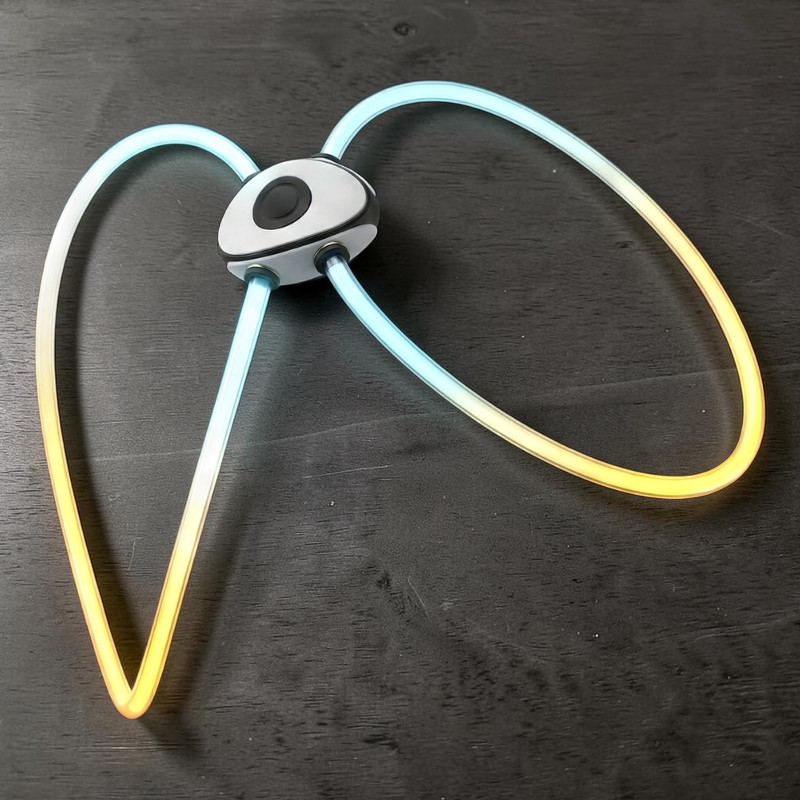 Harnais lumineux LED transparent pour chiens, visible sur fond sombre. Pièce centrale ovale grise avec bouton noir, avec quatre tubes transparents. Ils brillent du bleu clair à l'orange chaud et forment des boucles ouvertes.