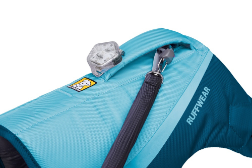 Gros plan sur le dos d'un harnais pour chien « Ruffwear » bleu clair, muni d'un éclairage de sécurité LED transparent sur le dessus. Une laisse noire est accrochée à une petite boucle/poche située sur le harnais. Le logo « Ruffwear » est visible.